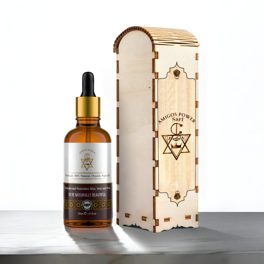 Amber glass dropper bottle labeled “Amigos Power Moroccan 100% Natural, Organic Argan Oil” standing next to an engraved wooden box featuring the brand’s logo. The back of the box displays detailed product information and ingredients in English and Greek.